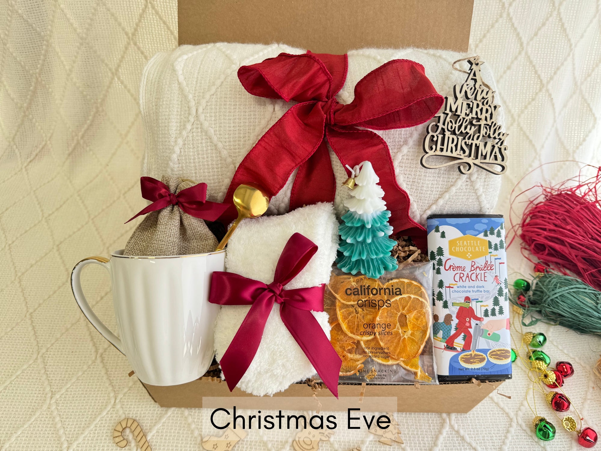 Christmas Eve Gift Box by Arinna Gifts featuring a throw blanket, festive socks, gourmet chocolate, coffee mug, elegant spoon, hot chocolate, candle, and Christmas ornaments.