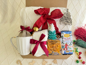 Christmas Eve Gift Box by Arinna Gifts featuring a throw blanket, festive socks, gourmet chocolate, coffee mug, elegant spoon, hot chocolate, candle, and Christmas ornaments.