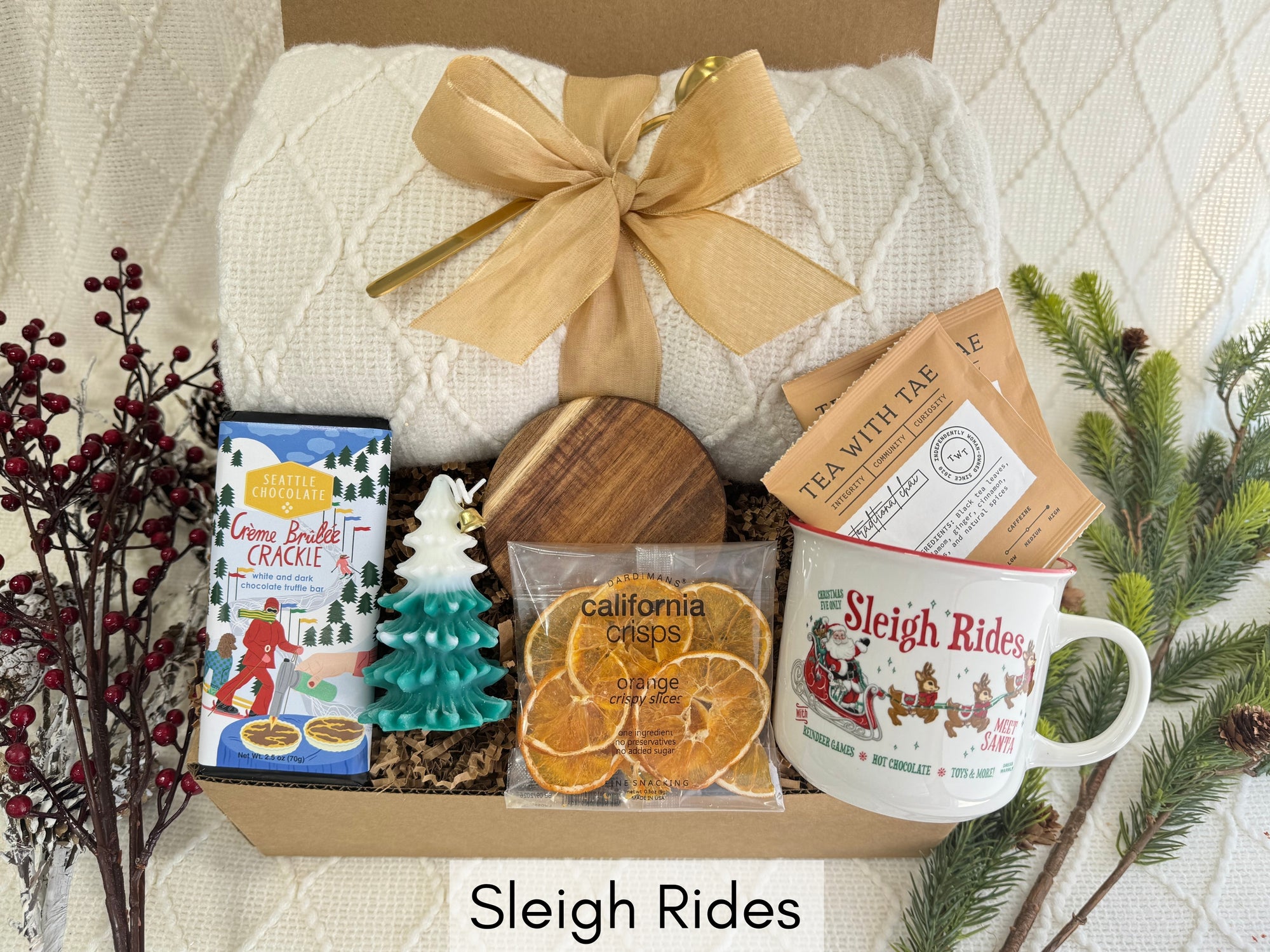 Sleigh Rides Gift Box featuring a throw blanket, gourmet chocolate, dried orange slices, Christmas coffee mug, golden coffee spoon, herbal tea, Christmas tree candle, and wooden coaster.