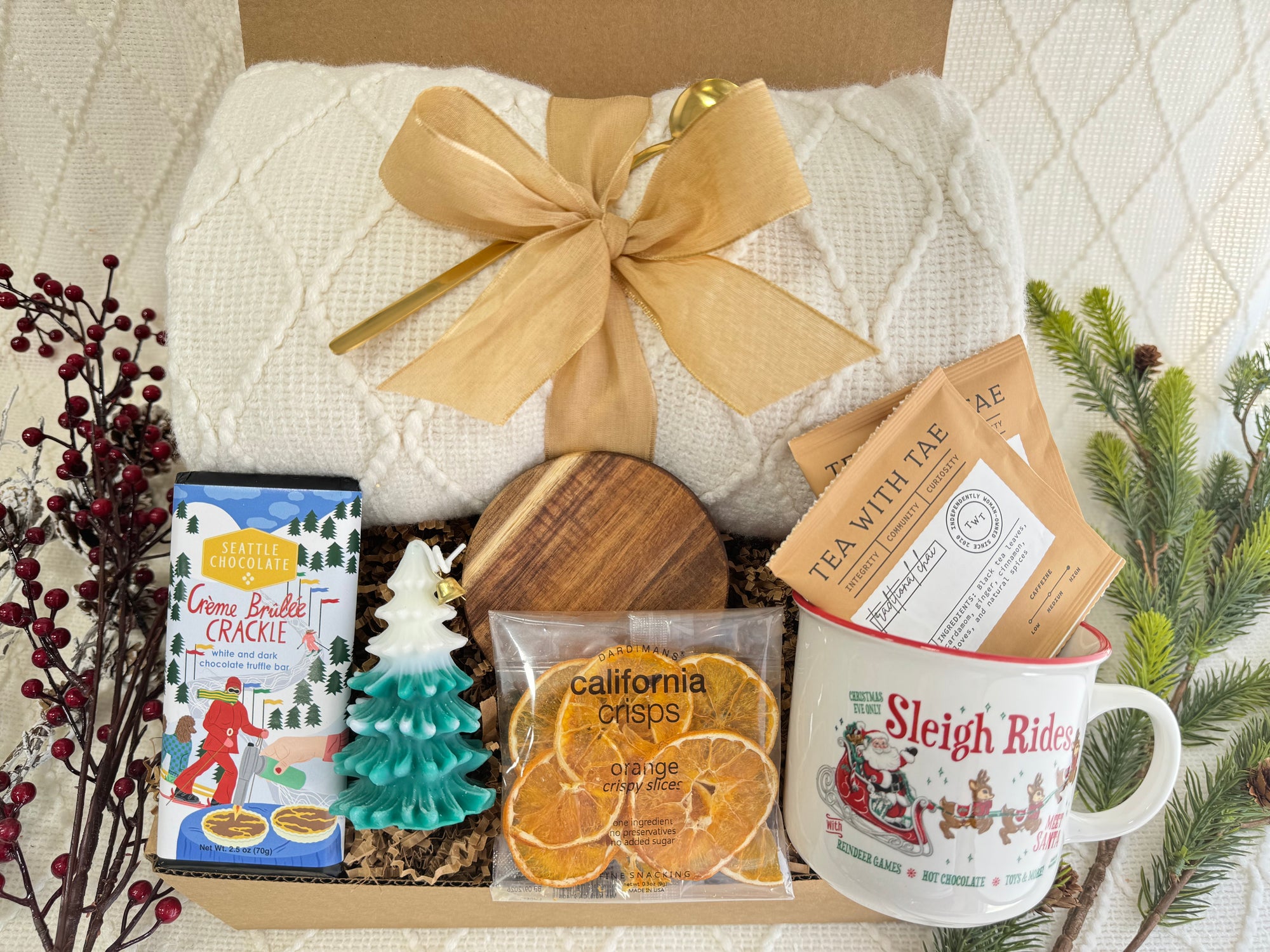 Sleigh Rides Gift Box featuring a throw blanket, gourmet chocolate, dried orange slices, Christmas coffee mug, golden coffee spoon, herbal tea, Christmas tree candle, and wooden coaster.