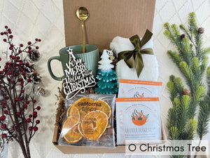 O Christmas Tree Gift Box featuring white fluffy socks, coffee mug, golden coffee spoon, dried orange slices, herbal tea, Christmas tree candle, and wooden Christmas ornament.