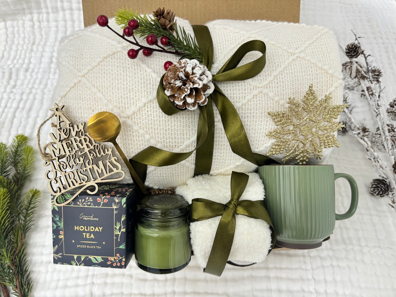 Holly-Jolly Gift Box featuring a throw blanket, fluffy socks, scent candle, coffee mug, golden coffee spoon, snowflake Christmas ornament, holiday tea, and wooden Christmas ornament.