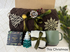 This Christmas Gift Box featuring a throw blanket, fluffy socks, coffee mug, Christmas tree candle, snowflake Christmas ornament, golden coffee spoon, holiday tea, and wooden Christmas ornament.