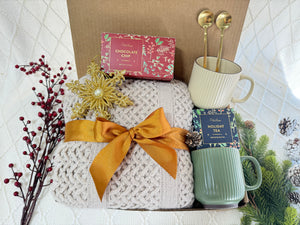 Home for Christmas Gift Box featuring a throw blanket, coffee mug, golden coffee spoon, holiday tea, gourmet cookie, and snowflake Christmas ornament.