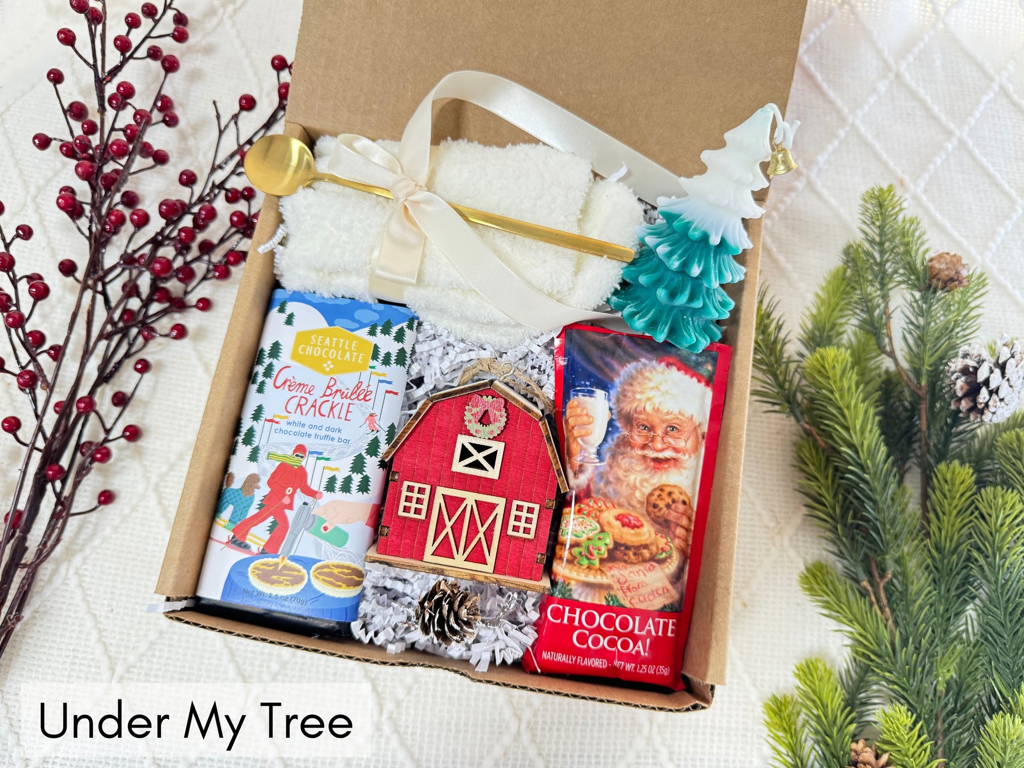 Under My Tree Gift Box featuring white fluffy socks, gourmet chocolate, wooden house ornament, Christmas tree candle, golden coffee spoon, and hot chocolate.