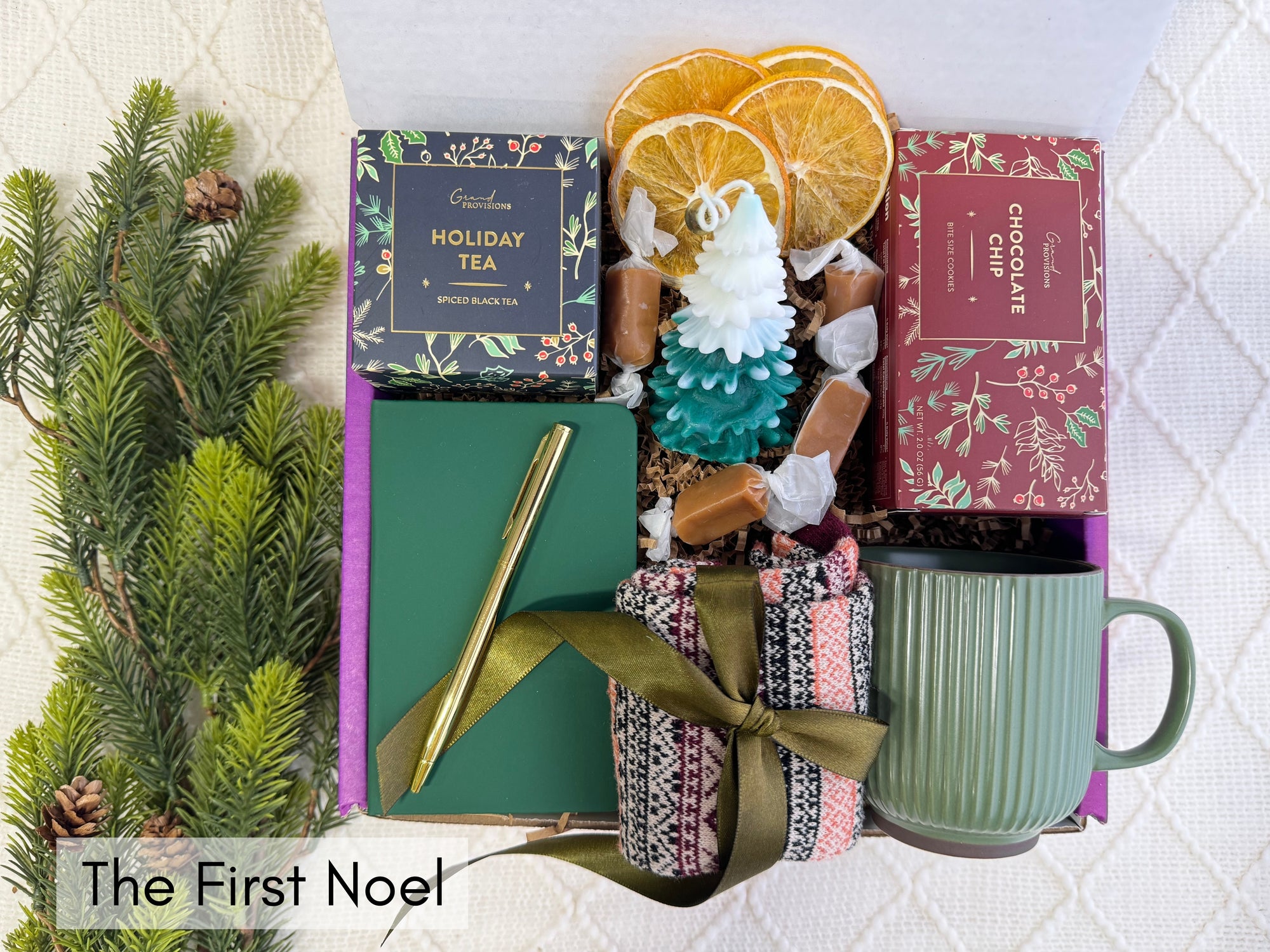 The First Noel Gift Box featuring a pocket notebook, Nordic socks, golden pen, holiday tea, Christmas tree candle, gourmet cookie, dried orange slices, coffee mug, and caramel candy.