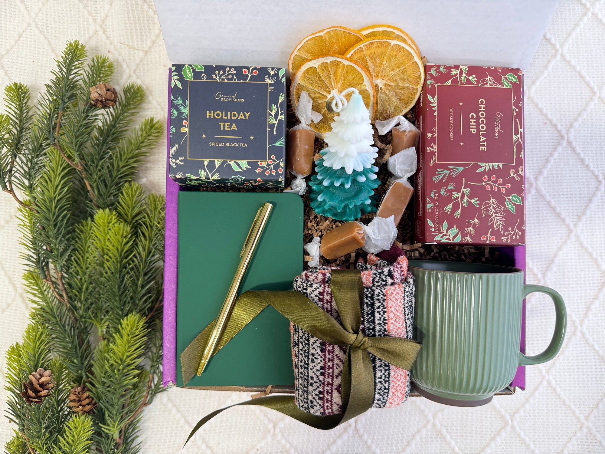 The First Noel Gift Box featuring a pocket notebook, Nordic socks, golden pen, holiday tea, Christmas tree candle, gourmet cookie, dried orange slices, coffee mug, and caramel candy.